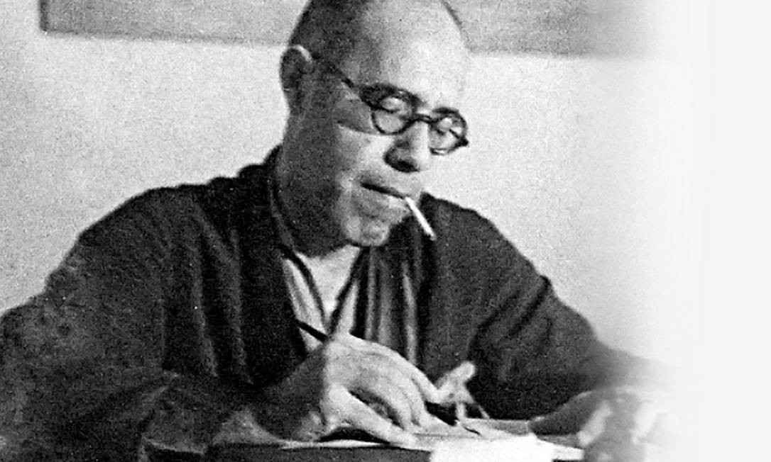 Imagem de Mário de Andrade, um homem pardo e calvo usando óculos de grau redondos e roupão. Mário fuma e tem diante de si um amontoado de papéis.
