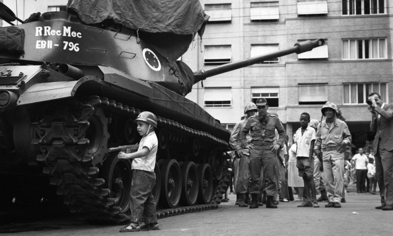 Uma criança usando capacete e portando uma arma de brinquedo é observada por soldados e civis enquanto brinca ao lado de um tanque do Exército Foto: Agência O Globo
