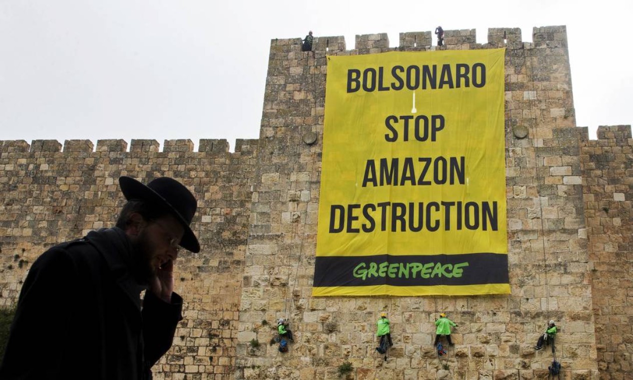 Homem ultra-ortodoxo caminha enquanto ativistas do Greenpeace penduram uma grande faixa nas muralhas da Cidade Velha de Jerusalém com uma mensagem para o presidente Bolsonaro em visita, em 1º de abril de 2019 Foto: OHAD ZWWINGENBERG / AFP