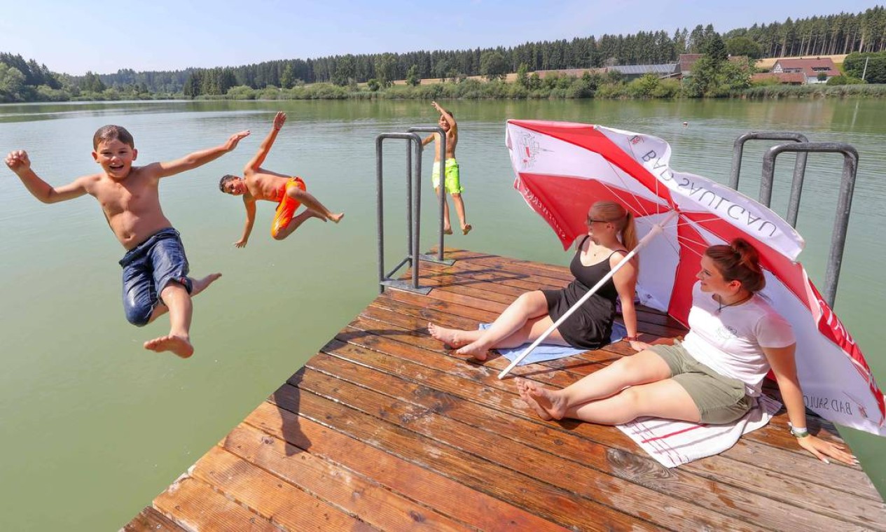 Duas mulheres sentadas sob guarda-sóis observam crianças pulando na água de um lago em Bad Saulgau, no sul da Alemanha, onde as temperaturas chegaram a 35°C nesta quinta-feira (25) Foto: THOMAS WARNACK / AFP