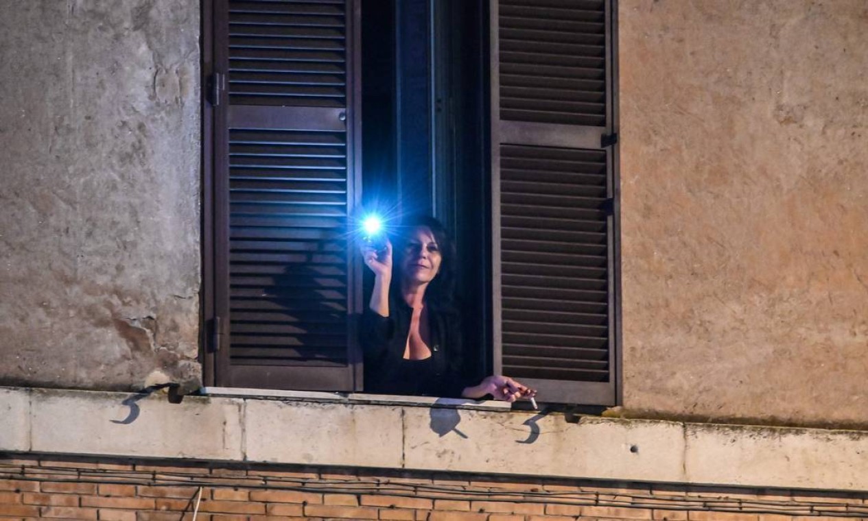 Mulher usa a lanterna de seu celular em um flashmob nas janelas organizado por italianos confinados Foto: ANDREAS SOLARO / AFP