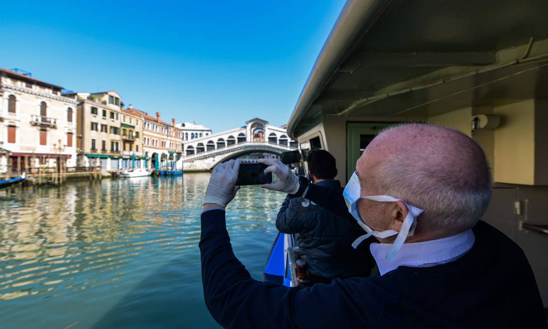 Homem usando máscara fotografa passageiros de uma balsa em Veneza: águas mais limpas após restrições a circulação de pessoas Foto: ANDREA PATTARO/AFP/18-3-2020