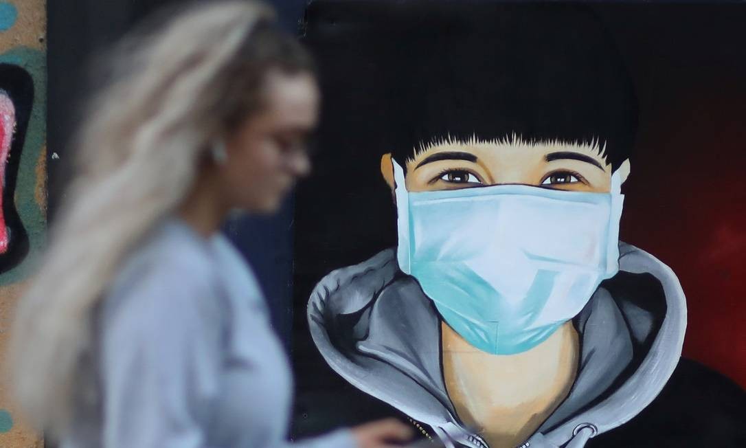 Em Sale, no Reino Unido, uma mulher confere o celular enquanto passa em frente a um grafite que mostra uma figura usando máscara cirúrgica, numa alusão às medidas adotadas contra a pandemia do novo coronavírus Foto: Phil Noble / Reuters