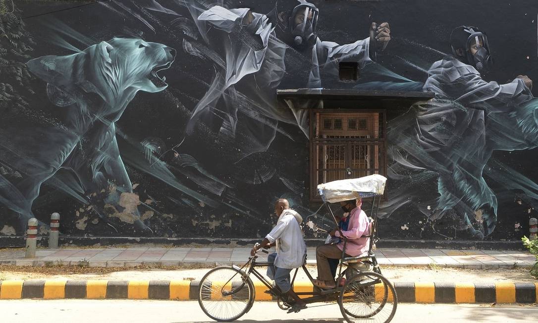 Riquixá carregando passageiros usando máscaras cirúrgicas passa em frente a um mural que retrata o combate ao novo coronavírus, no Lodhi Art District, em Nova Delhi, na Índia Foto: Sajjad Hussain / AFP