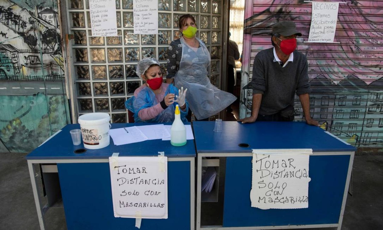 Voluntários esperam, ao lado das placas em que se lê "Manter distância. Somente com máscaras faciais", os residentes para um almoço solidário no bairro de La Pintana, Santiago, no Chile Foto: MARTIN BERNETTI / AFP