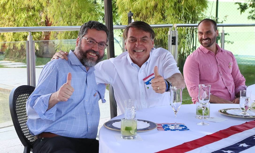Presidente Bolsonaro abraça o ministro das Relações Exteriores, Ernesto Araújo, se abraçam à mesa, ao lado do deputado federal e filho, Eduardo Bolsonaro Foto: Divulgação / Divulgação