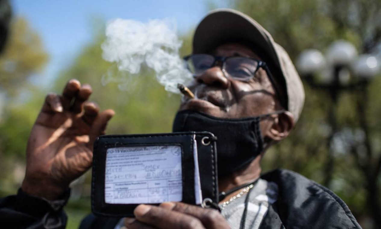 Homem fumando mostra seu cartão de vacinação enquanto ativistas oferecem erva para vacinados contra Covid-19 Foto: ANGELA WEISS / AFP