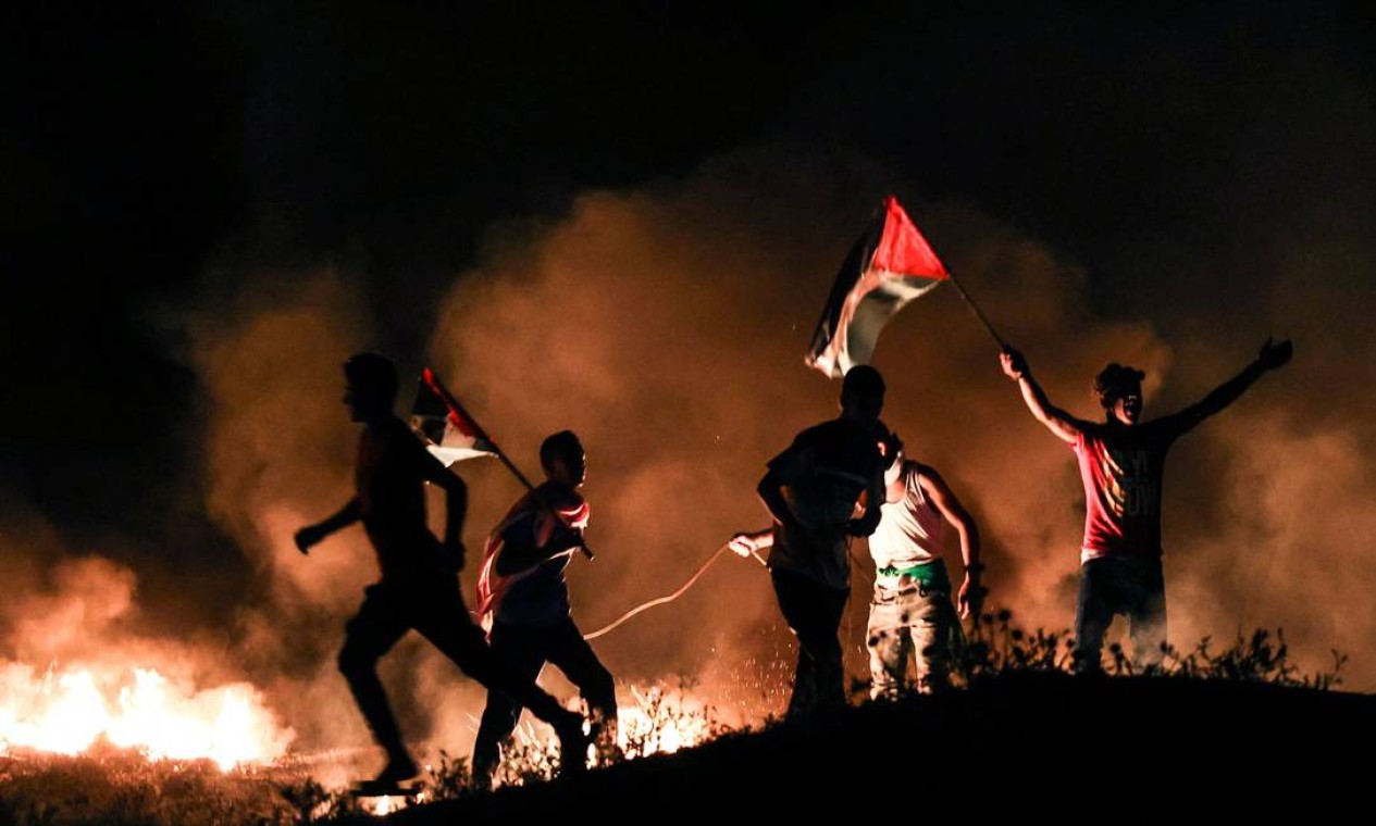 Manifestantes palestinos levantam bandeiras nacionais diante de fogueira de pneus ao leste da Cidade de Gaza perto da fronteira com Israel, em protesto contra a marcha das Bandeiras, movimento da extrema direita israelense Foto: MAHMUD HAMS / AFP