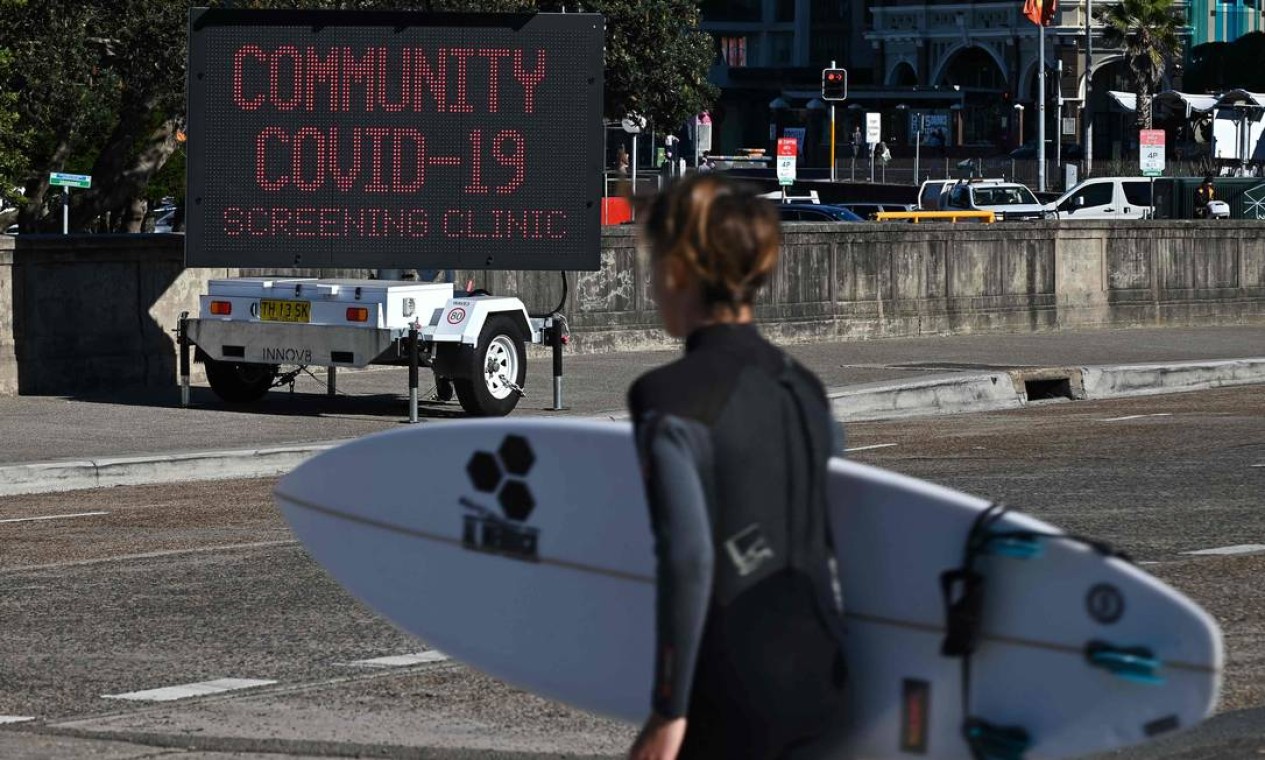 Surfista passa por uma placa para uma clínica de testes COVID-19, em Bondi Beach, Sydney Foto: STEVEN SAPHORE / AFP