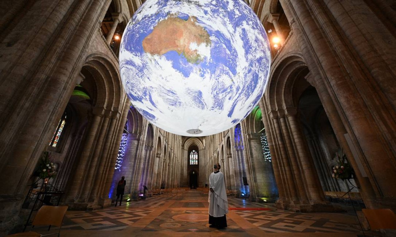 Membro do clero posa sob a instalação do artista britânico Luke Jerram 'Gaia', uma réplica de 7 metros do planeta Terra, suspensa na nave da Catedral de Ely em Ely, Cambridge Foto: OLI SCARFF / AFP
