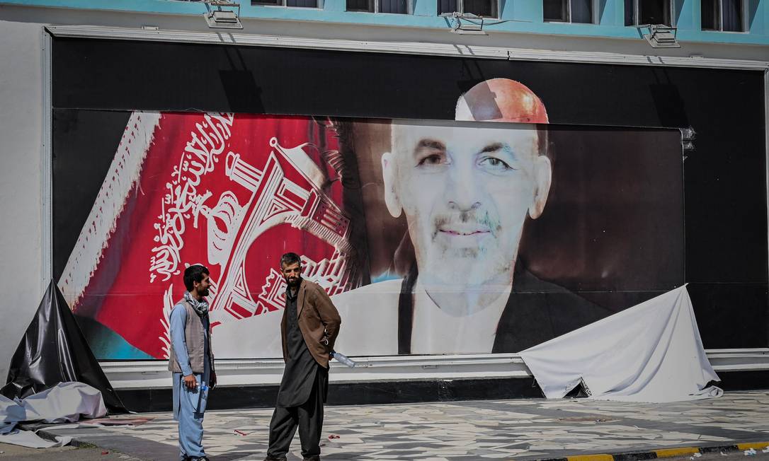 Homens conversam na frente de cartaz destruído com a imagem do ex-presidente Ashraf Ghani, em Cabul Foto: WAKIL KOHSAR / AFP