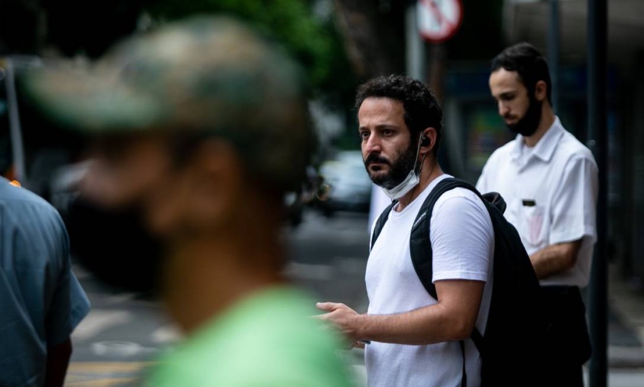 Homem usando máscara passa por outros dois, que estão com o acessório no queixo, à espera de um ônibus Foto: Brenno Carvalho / Agência O Globo