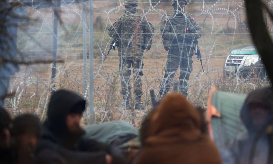 Imigrantes acampados do lado bielorrusso da fronteira observam mlilitares poloneses do lado oposto da cerca de arame farpado Foto: RAMIL NASIBULIN / AFP