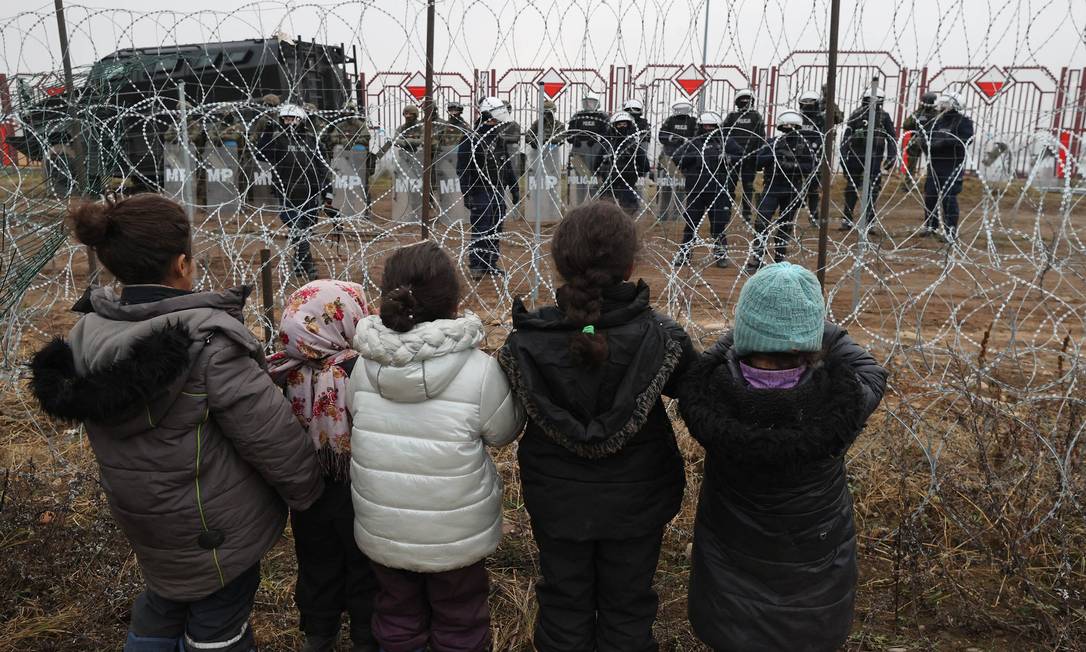 Crianças diante de arame farpado observam policiais no posto de fronteira de Bruzgi-Kuznica, entre a Bielorrússia e a Polônia Foto: MAXIM GUCHEK / AFP