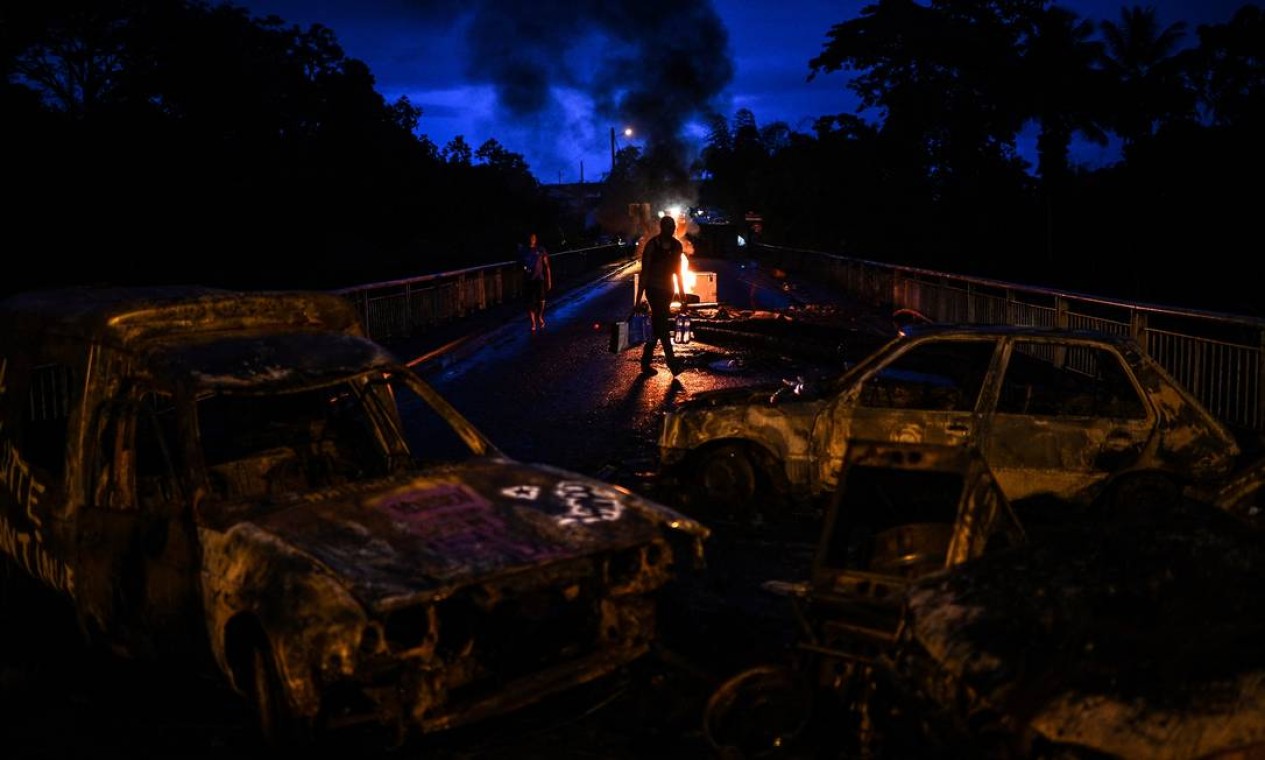 Homem carrega garrafas de água por um bloqueio de estrada em chamas feito de veículos queimados e destroços na localidade isolada de La Boucan, em Sainte-Rose, na ilha caribenha francesa de Guadalupe Foto: CHRISTOPHE ARCHAMBAULT / AFP