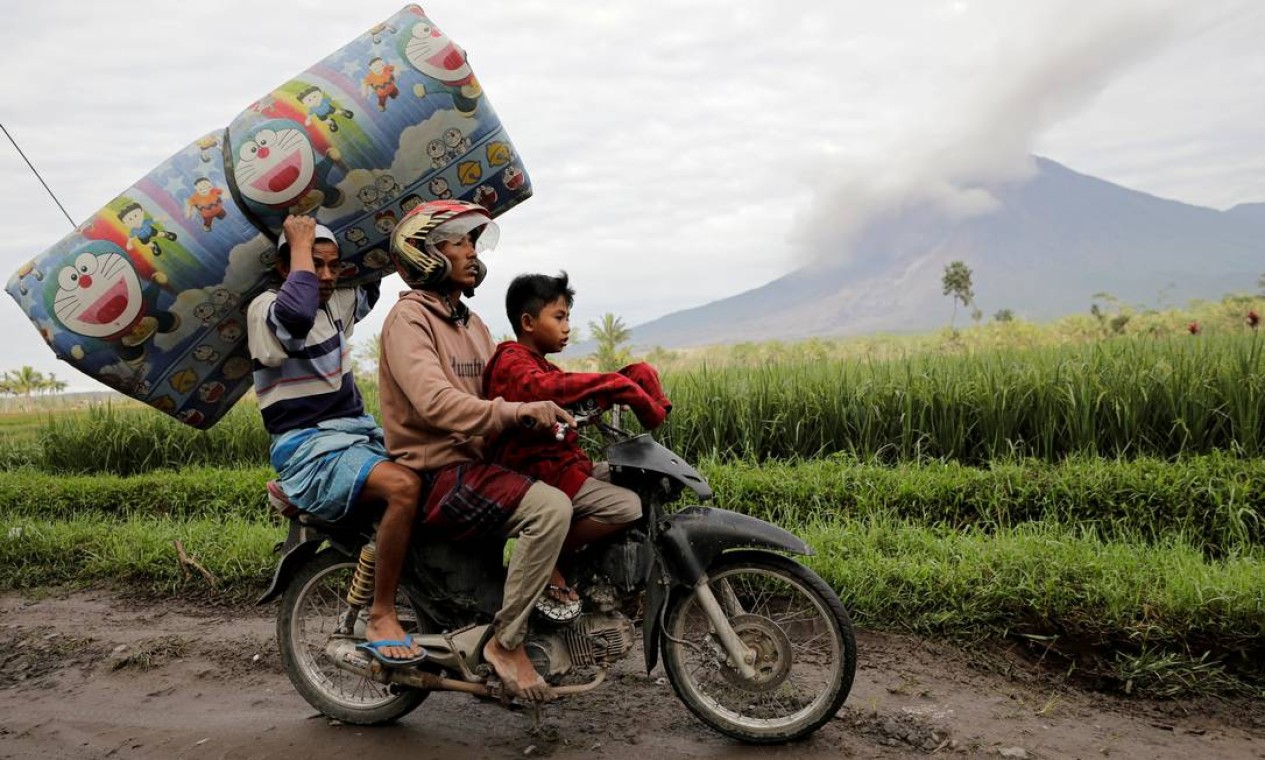 Moradores transportam um colchão em uma motocicleta enquanto o vulcão Monte Semeru continua a expelir cinzas e fumaça no fundo após uma erupção, em Gunung Sawur, Candipuro, Indonésia Foto: WILLY KURNIAWAN / REUTERS