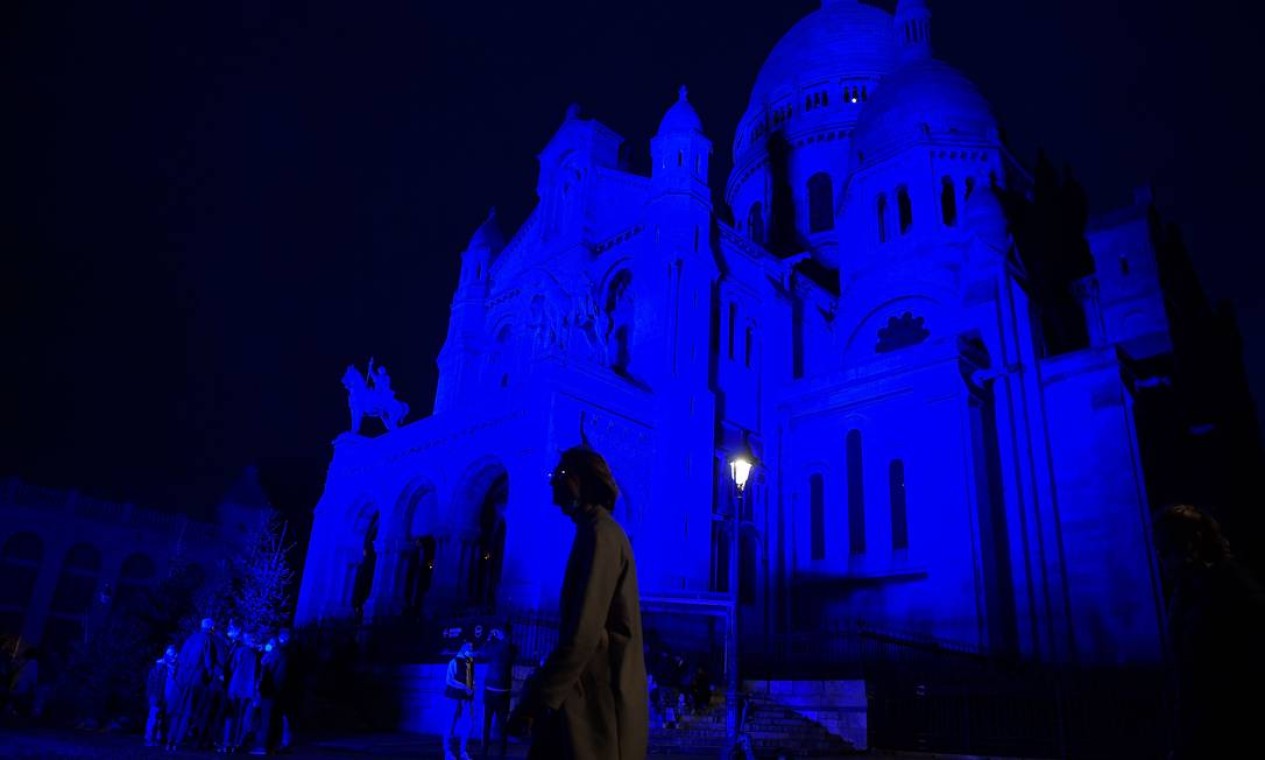 Pedestres caminham em frente à Basílica do Sagrado Coração, no topo da colina de Montmartre, em Paris, iluminada em azul Foto: JULIEN DE ROSA / AFP