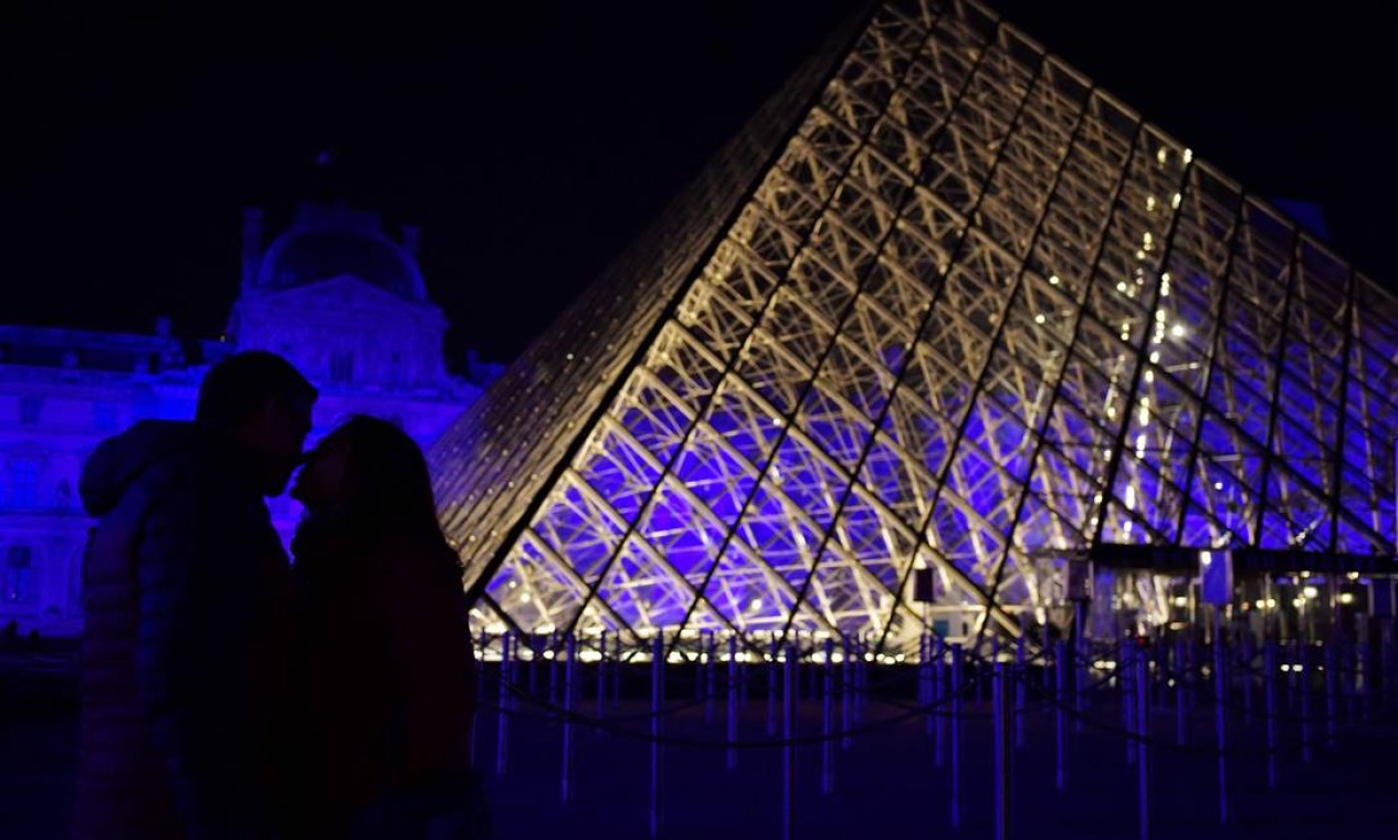 Casal se beija perto da Pirâmide na praça do Museu do Louvre, iluminada de azul Foto: JULIEN DE ROSA / AFP