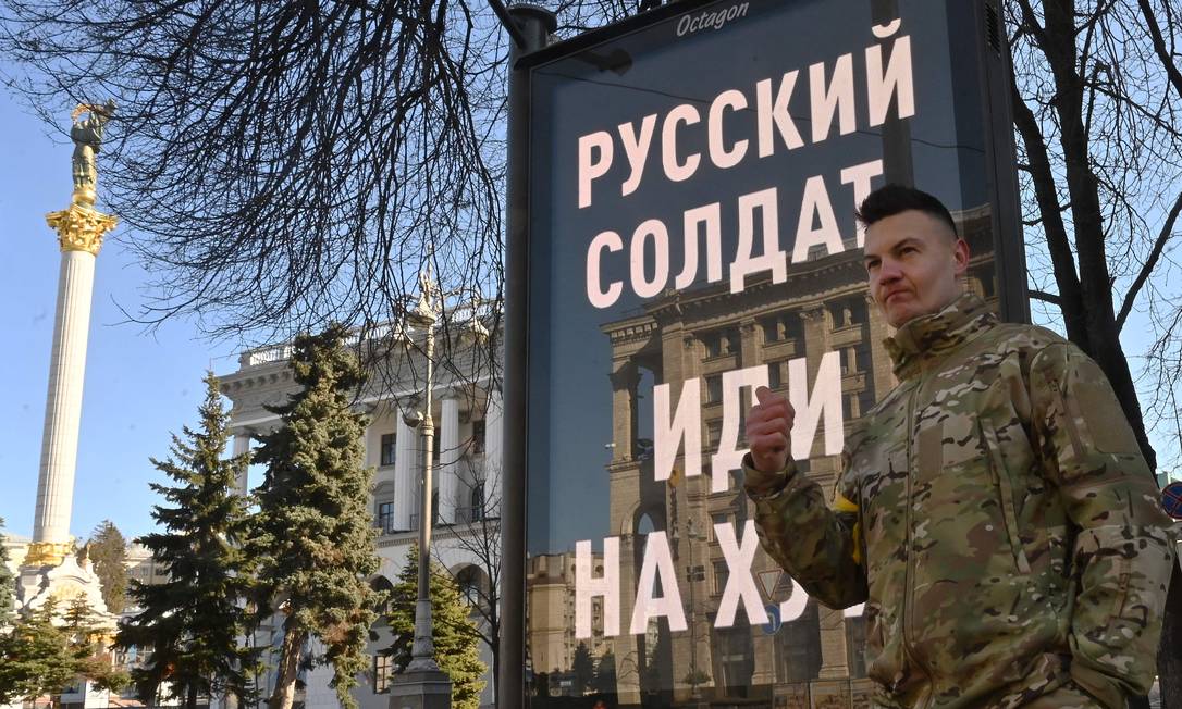Militar das Forças de Defesa Territorial da Ucrânia parado diante de cartaz que traz uma frase ofensiva aos russos: "Soldado russo, vá se f..." Foto: SERGEI SUPINSKY / AFP