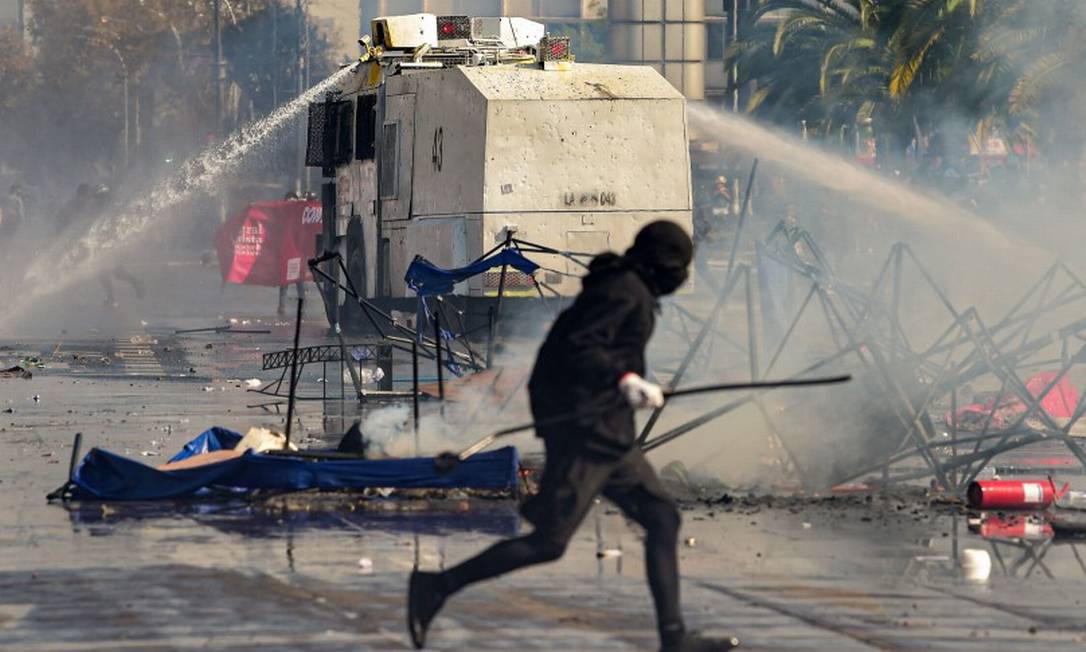 Batalhão de choque dispara canhão de água enquanto manifestante encapuzado corre em Santiago Foto: MARTIN BERNETTI / AFP