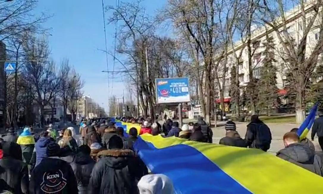 Captura de tela de imagens transmitidas ao vivo mostra pessoas carregando um banner nas cores da bandeira ucraniana, em Kherson, enquanto protestam em meio à invasão da Ucrânia pela Rússia Foto: Reprodução / Reuters / 13-3-2022