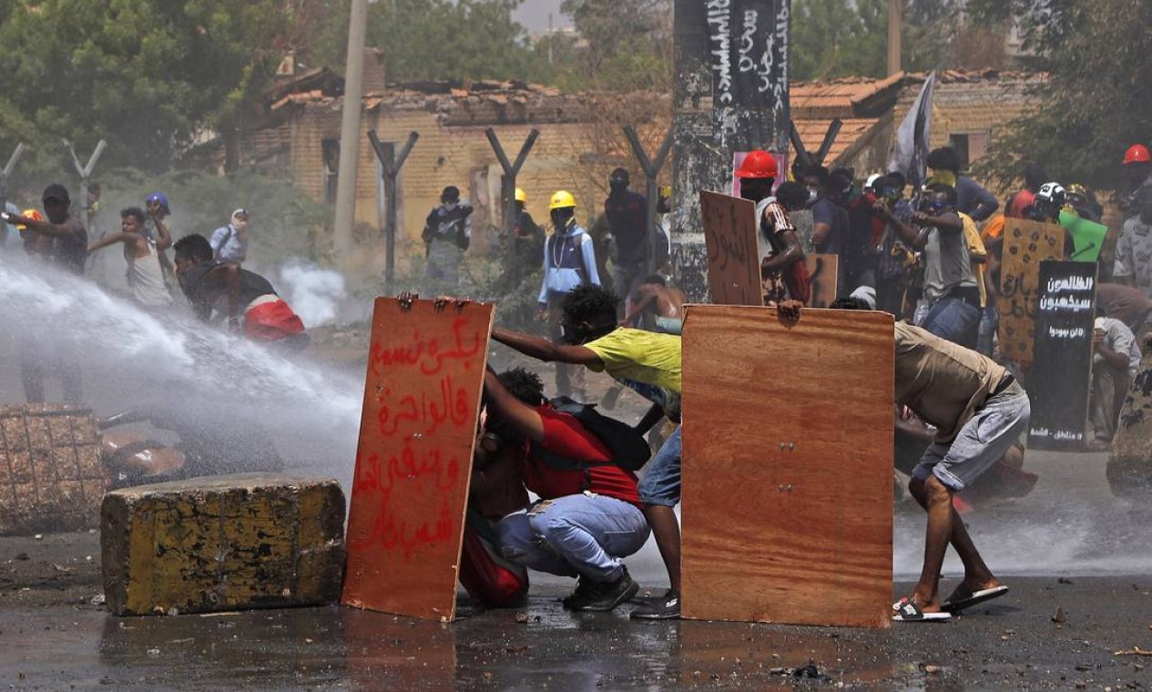 Manifestantes se protegem enquanto a tropa de choque tenta dispersá-los com canhões de água durante uma protesto contra o regime militar no centro da capital do Sudão, Cartum Foto: - / AFP