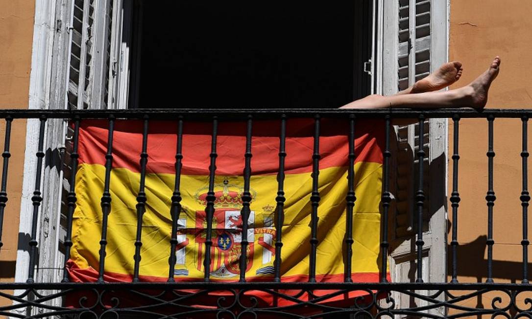 Mulher pega sol escondida atrás de uma bandeira da Espanha em sua varanda em Madri Foto: GABRIEL BOUYS / AFP